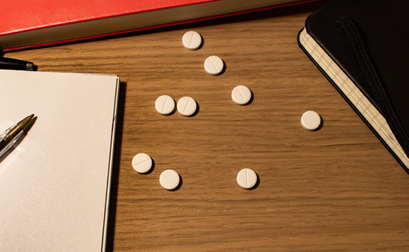 Pills scattered across a desk next to an open notebook with a pen on it.