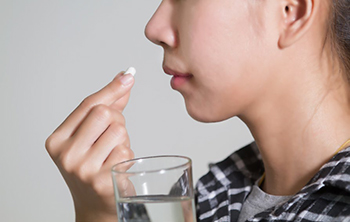 A young woman holds a pill close to her mouth in one hand and a glass of water in the other.