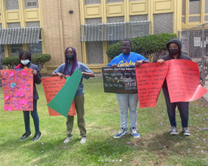 Four students hold large posters while standing on a lawn in front of a multistory brick building.