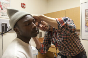 A Hispanic male physician uses an otoscope to look inside the ear of an African-American male student while standing in an exam room.