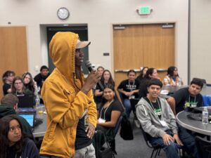 A teenaged boy wearing a yellow hoodie stands in a room full of seated youth while speaking into a microphone.